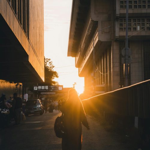 Person walking at sunset between buildings, capturing urban life and warm light in a bustling cityscape.